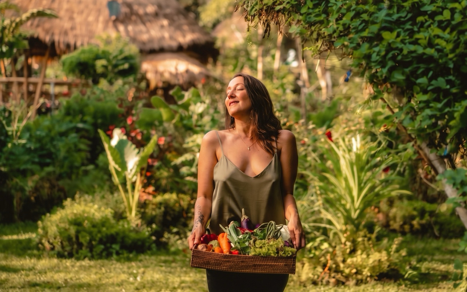 Jodie Louise standing in a sunlit tropical garden with her eyes closed, holding a woven basket of freshly picked vegetables