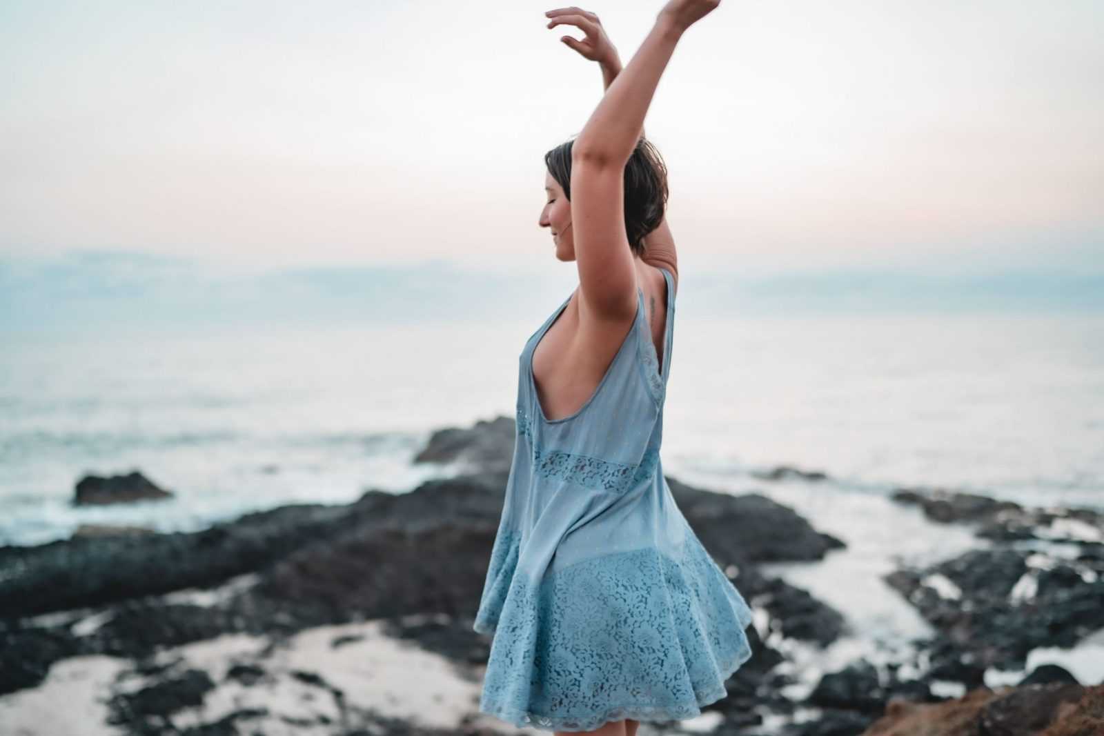 Jodie Louise standing by the sea at dusk with her arms raised, letting a feeling move through her
