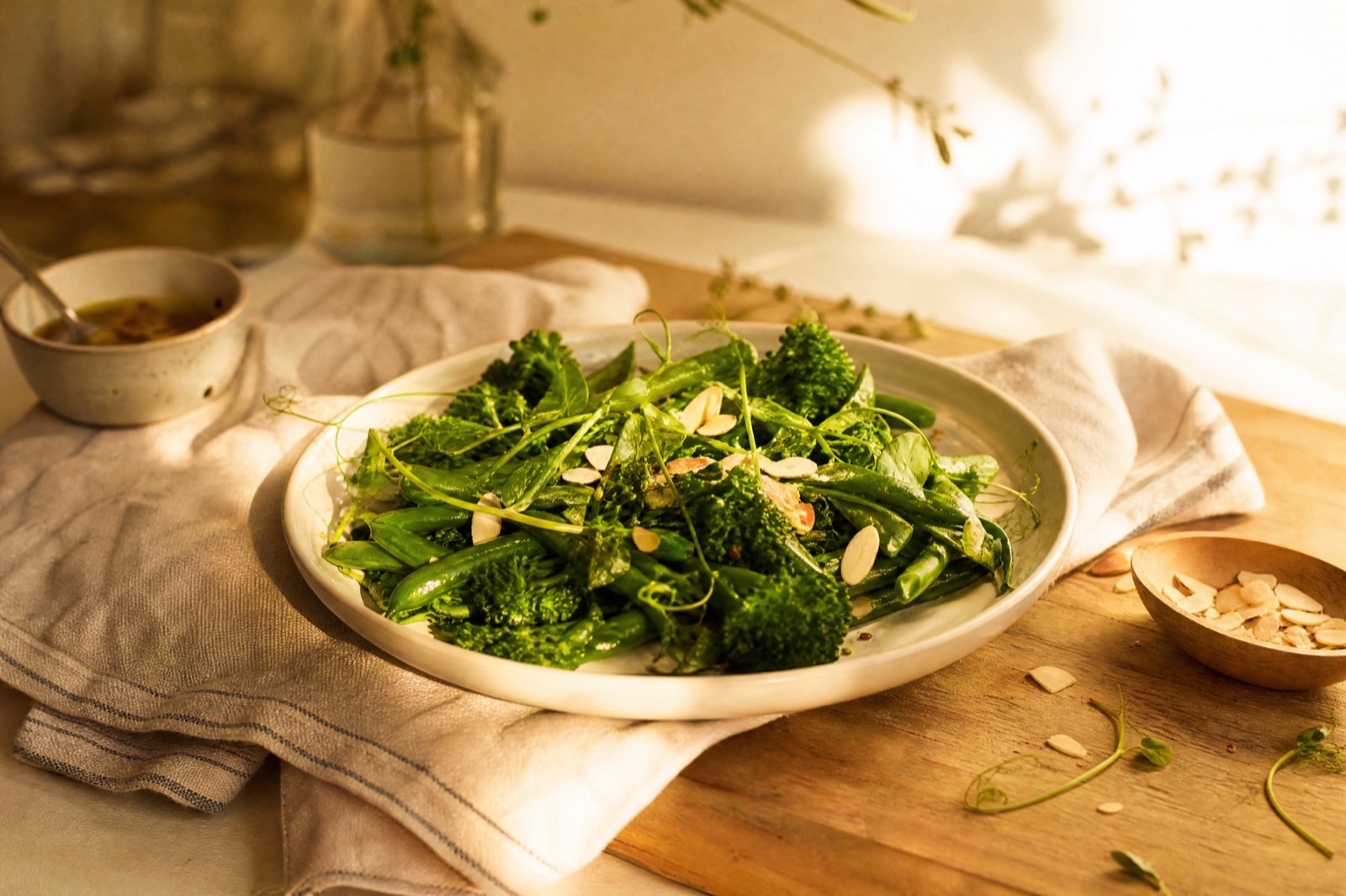A plate of steamed broccolini, snap peas and green beans scattered with toasted almonds, with a small bowl of dressing beside it