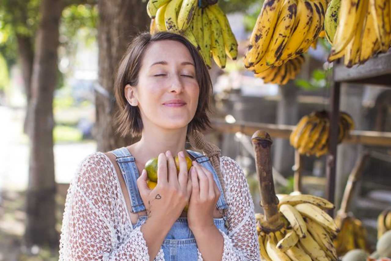 A woman standing at a fruit market with her eyes closed, holding a piece of fruit in both hands before eating it
