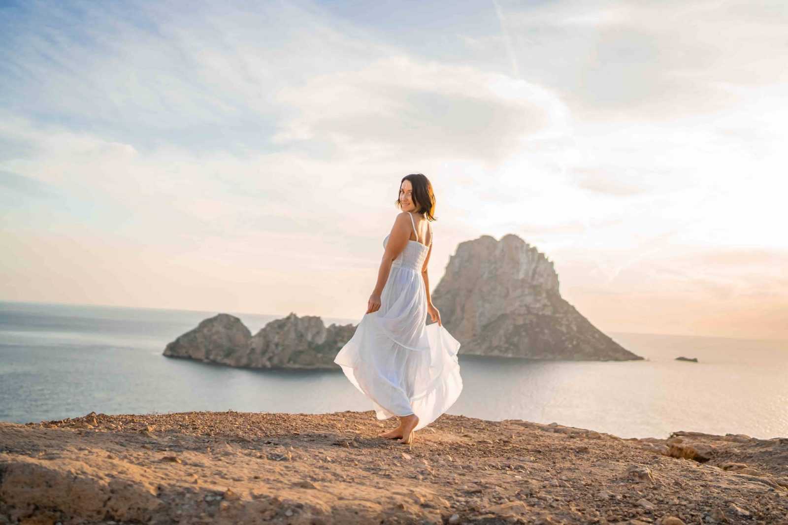 Jodie Louise in a white dress on the clifftop at sunset, with the rock of Es Vedra rising out of the sea behind her