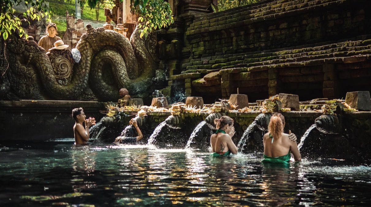 Bathers at the Tirta Empul purification spring in Bali, standing under the row of stone water spouts with offerings laid along the wall above them