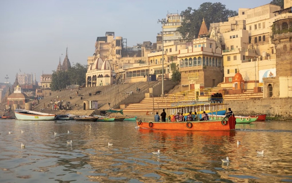 Boats on the Ganges at sunrise in Varanasi, with the stone ghats and temples of the old city rising behind the water