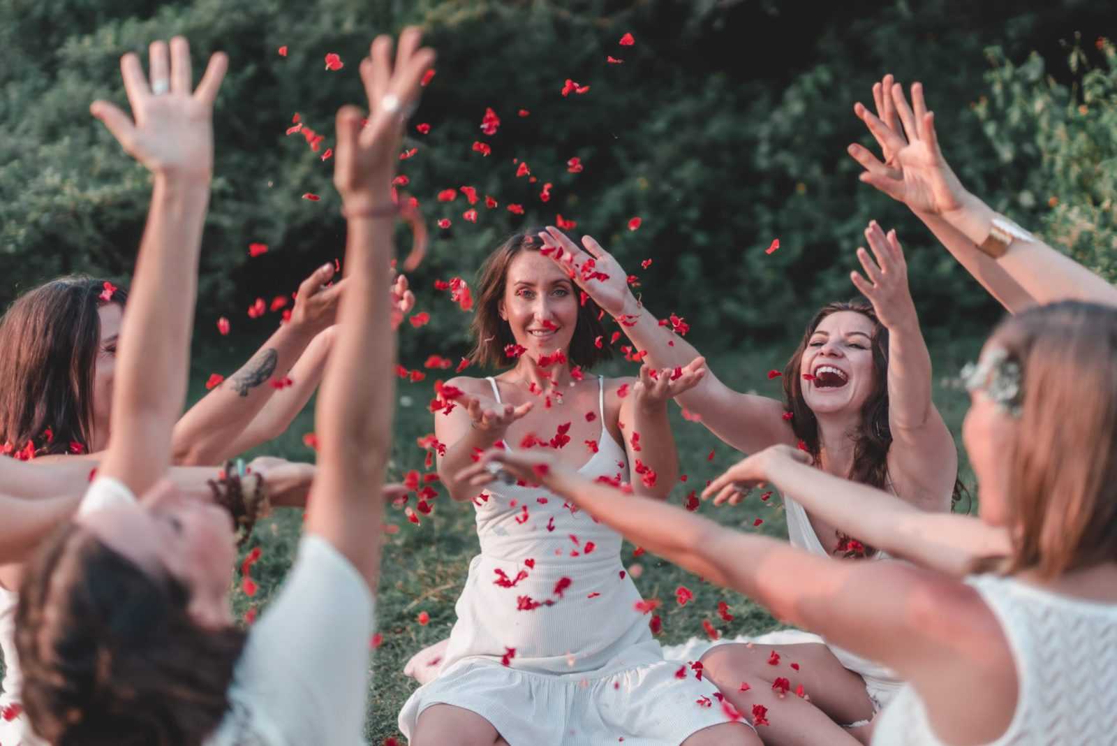 A circle of women sitting together on the grass, throwing rose petals into the air
