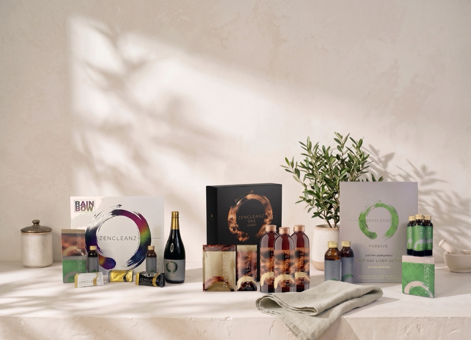 The three ZenCleanz kits, ONE, FORGIVE and the Rainbow Protocol, set out together on a pale plaster ledge beside an olive tree and folded linen, in soft morning light